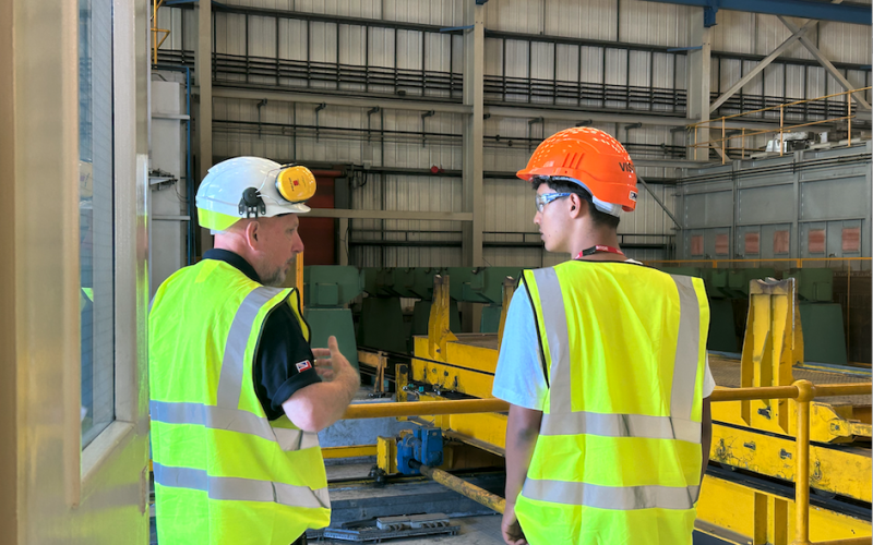Experienced factory worker in hard hard and hi-viz jacket talks to young worker, possibly on an apprenticeship, also in PPE